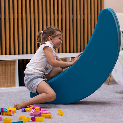 A girl smiling, rocking on a blue banana shaped foam rocking toy